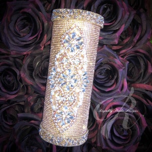 May include: A gold vase decorated with rhinestones and pearls, surrounded by dark red roses. The vase has a tall, cylindrical shape and is embellished with a diamond-shaped pattern of crystals.