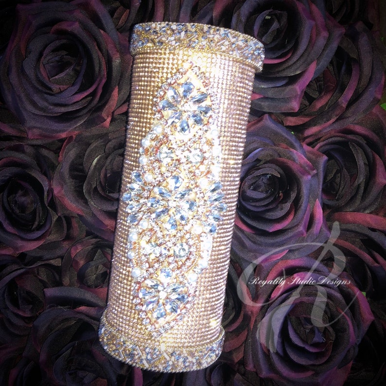 May include: A gold vase decorated with rhinestones and pearls, surrounded by dark red roses. The vase has a tall, cylindrical shape and is embellished with a diamond-shaped pattern of crystals.