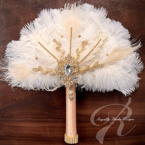 May include: A white and ivory feather bridal bouquet with a large crystal and gold rhinestone embellishment. The bouquet has a blush pink satin handle with a gold rhinestone band.