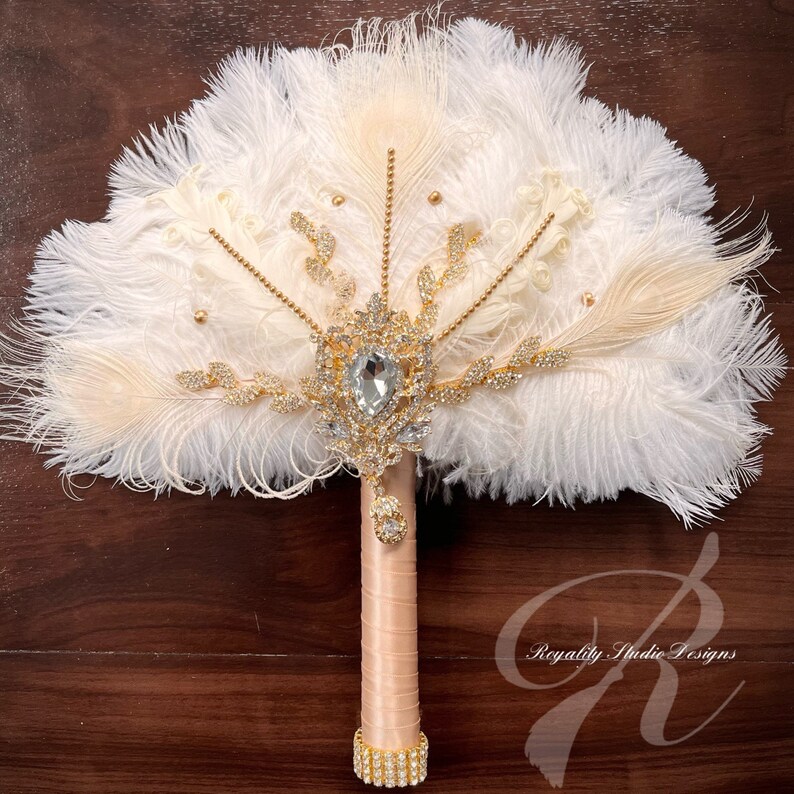 May include: A white and ivory feather bridal bouquet with a large crystal and gold rhinestone embellishment. The bouquet has a blush pink satin handle with a gold rhinestone band.