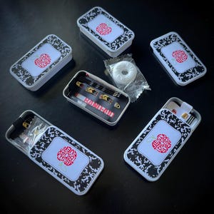 May include: Several small, rectangular metal tins with a white lid featuring a red apple design and the text "Best Teacher Ever". The tins are open, revealing small sewing kits, safety pins, and a wrapped mint.