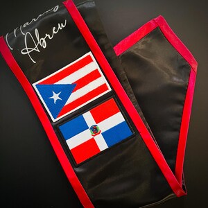 May include: Black satin sash with red trim and two embroidered patches. The patches feature the flags of Puerto Rico and the Dominican Republic. The sash is personalized with the name "Maria Abreu".