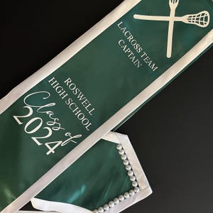 May include: Green and white graduation sash with the text "Class of 2024" and "Roswell High School". The sash also has the text "Lacrosse Team Captain" and a white lacrosse stick graphic.