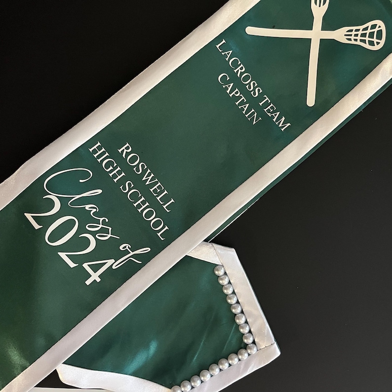 May include: Green and white graduation sash with the text "Class of 2024" and "Roswell High School". The sash also has the text "Lacrosse Team Captain" and a white lacrosse stick graphic.