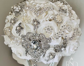 May include: A white bridal bouquet made of silk roses and adorned with a variety of silver brooches and pearl accents.