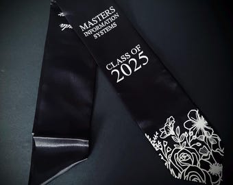Puede incluir: Banda de graduación de satén negro con texto blanco que dice "MASTERS INFORMATION SYSTEMS" y "CLASS OF 2025". El borde inferior presenta un diseño floral blanco. La banda se muestra sobre un fondo oscuro.