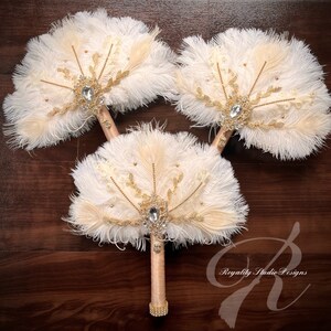 May include: Three white and beige feather fan wedding bouquets with gold accents and rhinestone embellishments. The bouquets have a vintage style.