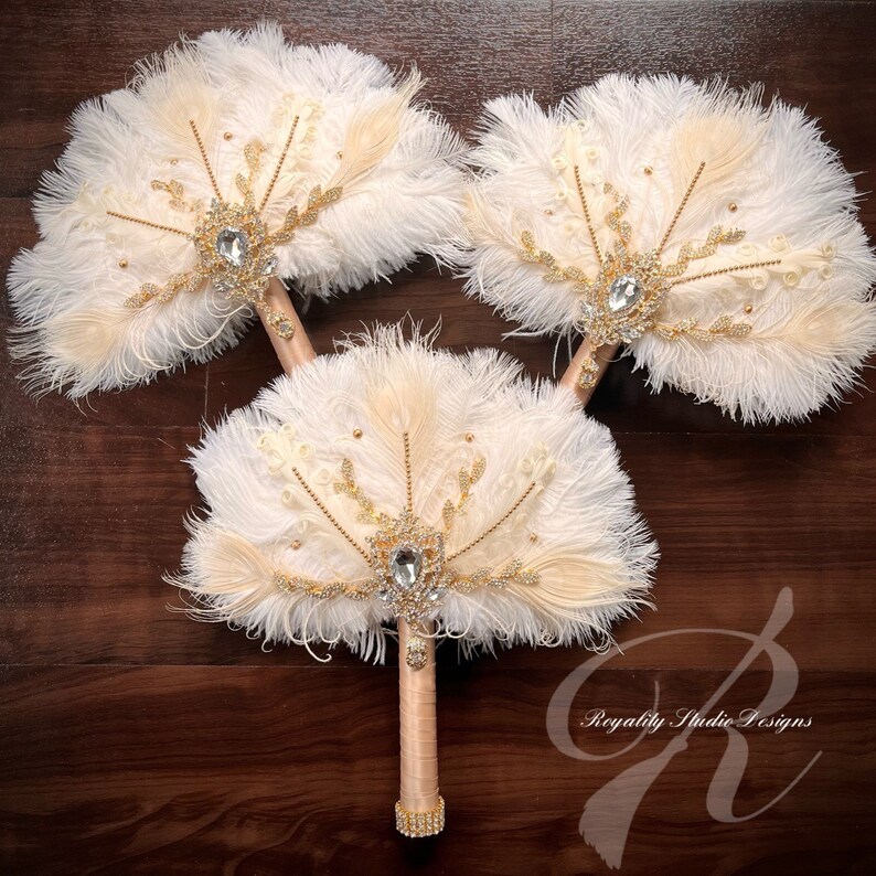 May include: Three white and beige feather fan wedding bouquets with gold accents and rhinestone embellishments. The bouquets have a vintage style.