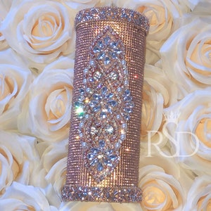 May include: A rose gold tumbler with a decorative design of clear rhinestones and white pearls. The tumbler is surrounded by white roses.