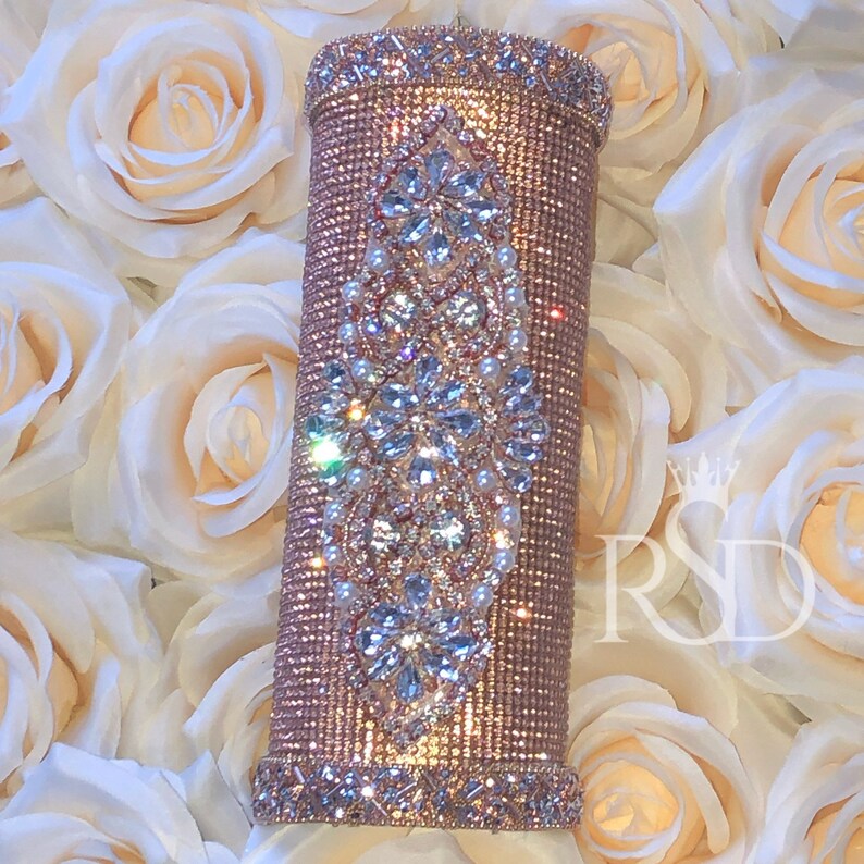 May include: A rose gold tumbler with a decorative design of clear rhinestones and white pearls. The tumbler is surrounded by white roses.