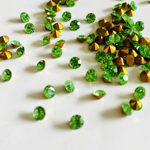 Puede incluir: Una dispersión de piedras de cristal verde con engastes dorados sobre una superficie blanca.