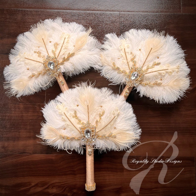 May include: Three white feather fans with gold accents and crystal embellishments. The fans are arranged in a triangular shape on a wooden surface. The text "Royalty Studio Designs" is visible in the bottom right corner of the image.
