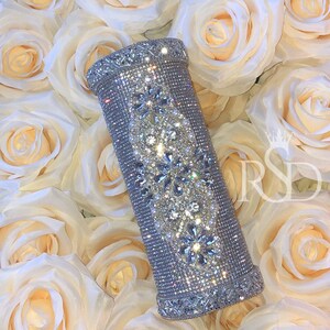 May include: A silver cylinder-shaped clutch purse covered in sparkling rhinestones and pearls. The purse has a decorative design of floral patterns.