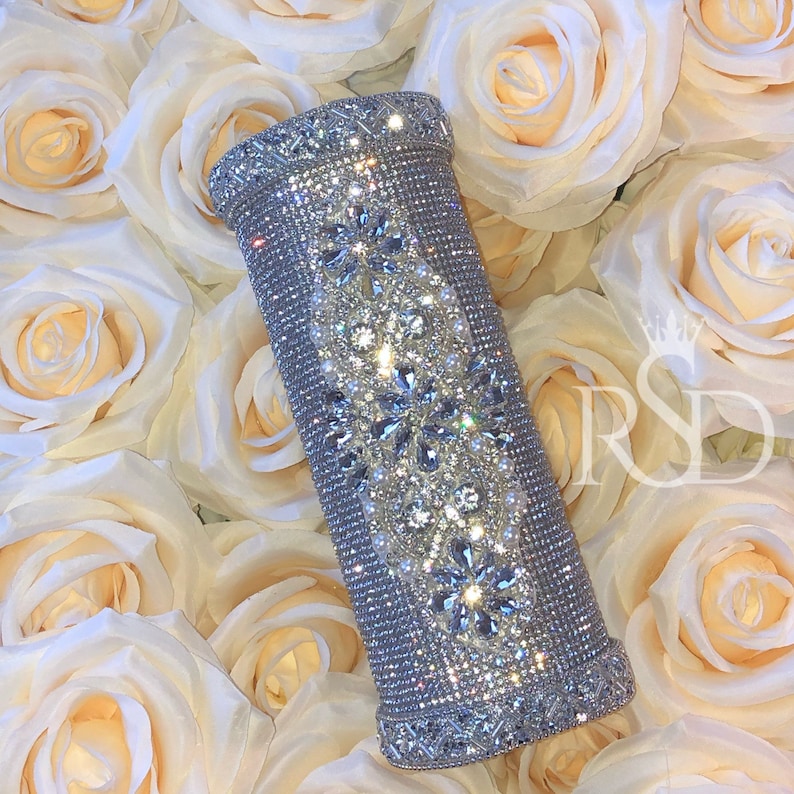May include: A silver cylinder-shaped clutch purse covered in sparkling rhinestones and pearls. The purse has a decorative design of floral patterns.