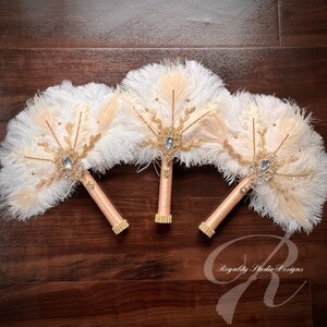 May include: Three white and beige feather fan bouquets with gold accents and jeweled brooches. The bouquets have peach satin stems and are arranged on a wooden surface.