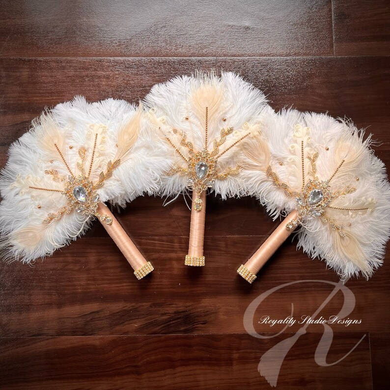 May include: Three white and beige feather fan bouquets with gold accents and jeweled brooches. The bouquets have peach satin stems and are arranged on a wooden surface.