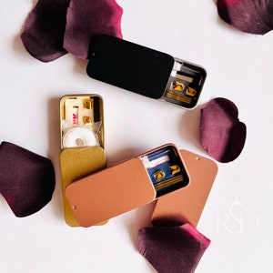 Puede incluir: Tres pequeñas latas de metal con tapas de diferentes colores. Las latas están abiertas y revelan varios artículos en su interior, incluidos vendajes, agujas de coser e hilo. Las latas están sobre una superficie blanca con algunos pétalos de rosa burdeos.