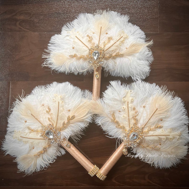 May include: Three white and beige feather fans with gold accents and jeweled brooches. The fans are tied together with a gold ribbon.