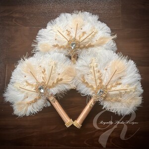 May include: Three white and beige feather fans with gold accents and crystal embellishments. The fans are arranged in a triangle shape and have gold handles.