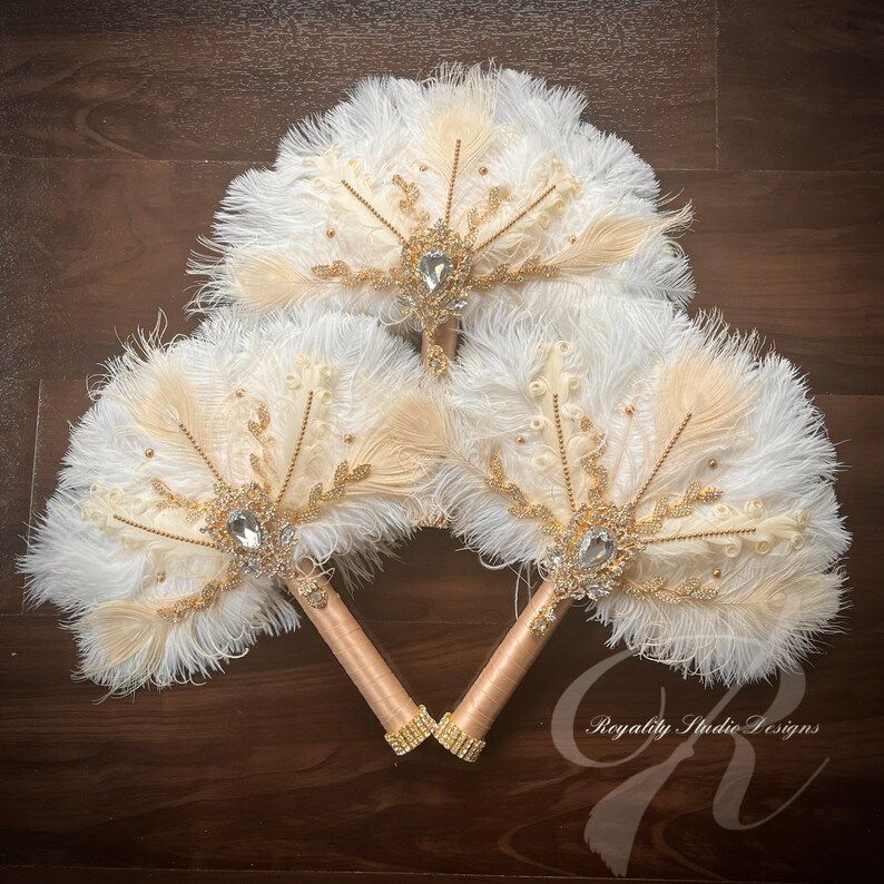 May include: Three white and beige feather fans with gold accents and crystal embellishments. The fans are arranged in a triangle shape and have gold handles.