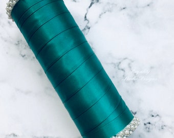 May include: Teal satin ribbon wrapped around a cylinder with a pearl trim. The cylinder is on a white marble surface.