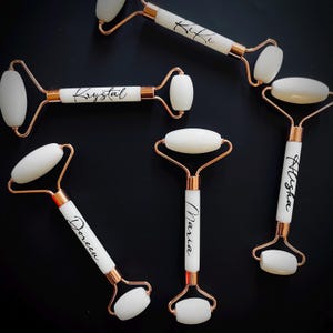 May include: Several white and rose gold facial rollers with names printed on the handles. The rollers have a white stone and are set against a black background. The names include Kiki, Krystal, Alysha, Maria, and Doreen.