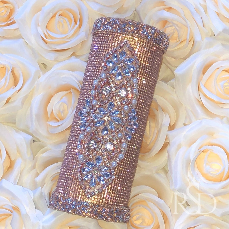 May include: A rose gold clutch purse covered in rhinestones and pearls. The clutch is decorated with a large, ornate rhinestone design.