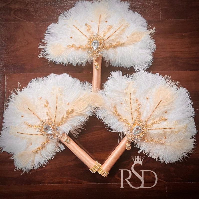 May include: Three white and peach feather fans with gold accents and jeweled embellishments. The fans are arranged in a triangle shape on a wooden surface.