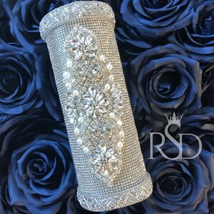 May include: A silver cylindrical vase decorated with rhinestones and pearls. The vase is surrounded by dark blue fabric roses.