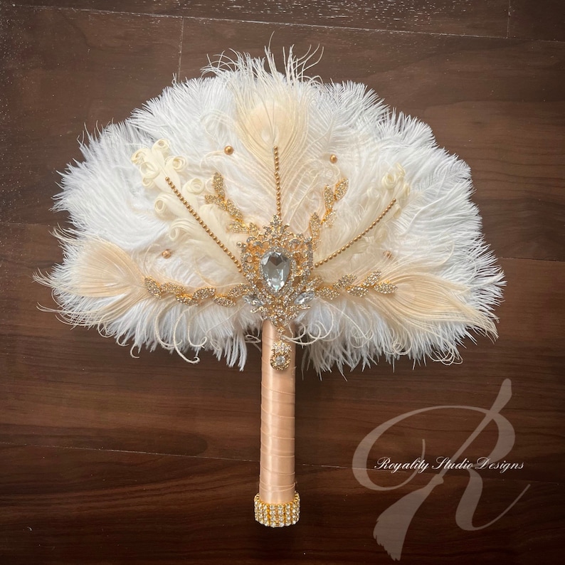 May include: A white and ivory feather fan bridal bouquet with a gold and crystal brooch. The handle is wrapped in ivory satin and has a gold rhinestone band.