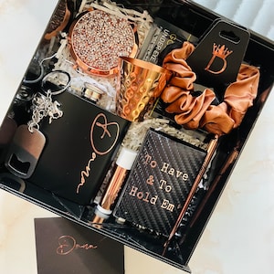 May include: A black gift box filled with various items, including a black flask with a rose gold heart and the word "home" engraved on it, a copper shot glass, a black card with the text "To Have & To Hold Em", a rose gold lip balm, a brown hair scrunchie, a rose gold glitter coaster, and a black key chain with a bottle opener.