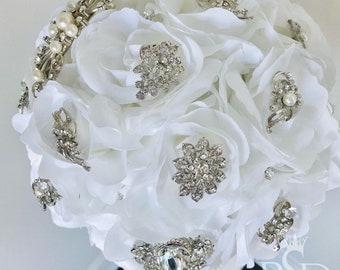 May include: A white silk flower bouquet decorated with vintage rhinestone brooches and pearl accents. The bouquet is perfect for a wedding or special occasion.