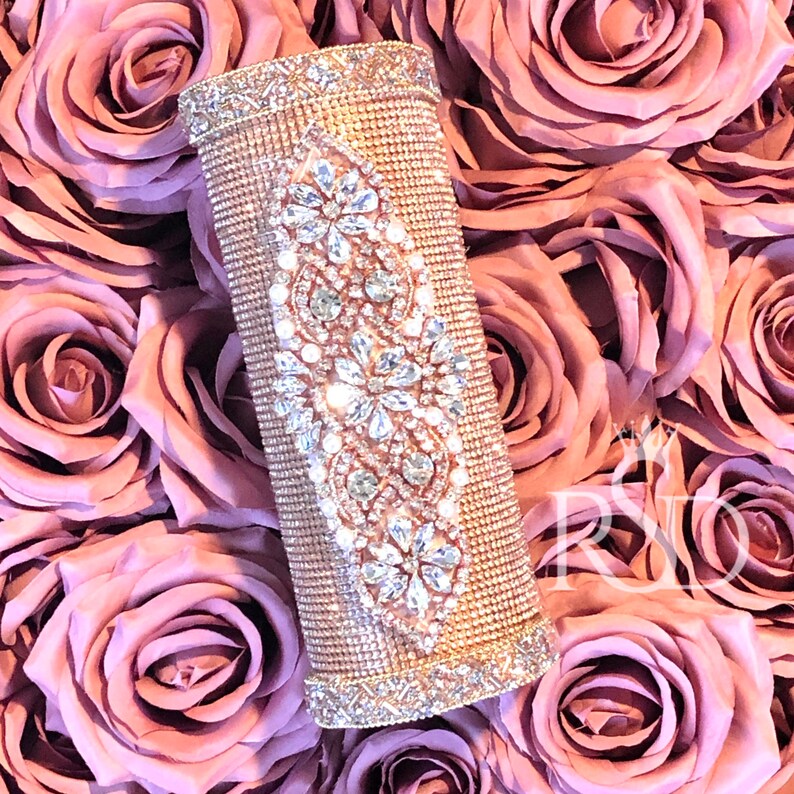 May include: A gold clutch purse with a diamond-shaped design covered in rhinestones. The purse is surrounded by pink roses.