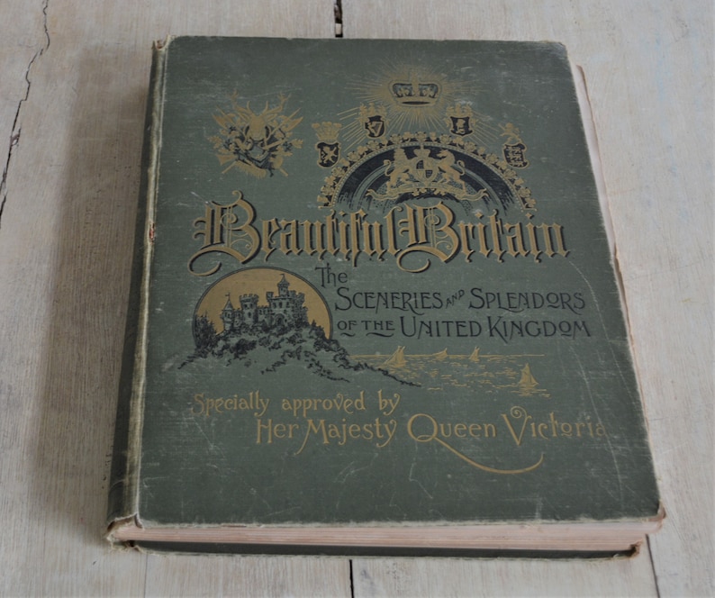 Op de afbeelding: Een groen hardback boek getiteld "Beautiful Britain: The Sceneries and Splendors of the United Kingdom" met gouden letters en illustraties. Het boek is versierd met een kroon en andere koninklijke symbolen. De tekst "Specially approved by Her Majesty Queen Victoria" staat op de omslag gedrukt.