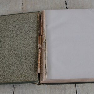 Op de afbeelding: Een oud, versleten boek met een groene, gepatroneerde omslag en een blanco witte pagina die open ligt. Het boek ligt open om de rug te laten zien, die beschadigd en versleten is.