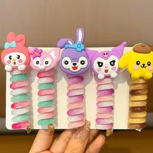 May include: A collection of five vibrant hair clips, each adorned with a cartoon character. The clips have a spiral design in pink, green, and brown. Characters include a pink bunny, a pink cat, a purple bunny, a purple cat, and a yellow dog.