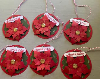 May include: Six handmade Christmas ornaments. Each ornament features a red poinsettia flower with a gold glitter center, set on a red circle with green scalloped edges. A white tag with "Merriest Wishes" is attached with twine.