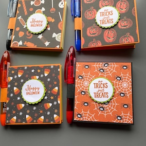 May include: Four Halloween themed notepad sets with pens attached. Each notepad has a different design: candy corn, pumpkins, spiders, and candy canes. The notepads have the text "Happy Halloween" and "No Tricks Just Treats" on them.