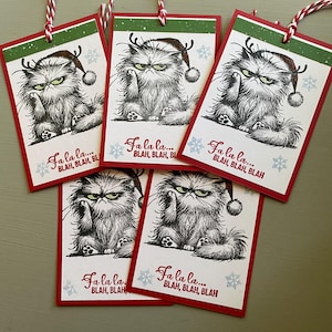 May include: A collection of Christmas gift tags featuring a grumpy cat illustration wearing a Santa hat with antlers. The tags have a red border, green accents, and the text "Fa la la... BLAH, BLAH, BLAH". Each tag is strung with red and white twine.