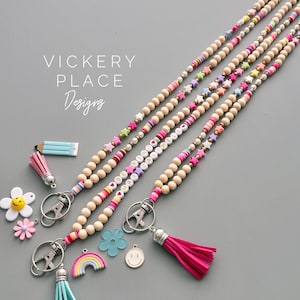 May include: Wooden bead lanyards with colorful accents and charms. The lanyards feature wooden beads, star-shaped beads, and letter beads spelling out words. Charms include a pencil, flowers, a rainbow, and a smiley face.