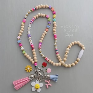 May include: A beaded lanyard with wooden beads, colorful star and striped beads, and the word "BE KIND" spelled out. It includes a pink tassel, a blue tassel, and various charms, including a flower, apple, and balloon dog.