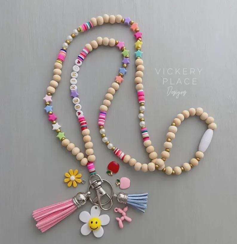 May include: A beaded lanyard with wooden beads, colorful star and striped beads, and the word "BE KIND" spelled out. It includes a pink tassel, a blue tassel, and various charms, including a flower, apple, and balloon dog.