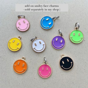 May include: A collection of colorful smiley face charms in various colors, including pink, blue, yellow, white, purple, green, orange, and black. Each charm has a gold-colored border and a silver jump ring for attachment. The text "add on smiley face charms ~ sold separately in my shop :)" is visible.