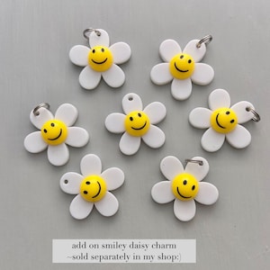May include: Seven white daisy charms with yellow smiley faces at the center. Each charm has a silver-colored ring attached at the top. The text at the bottom reads "add on smiley daisy charm ~sold separately in my shop:)".
