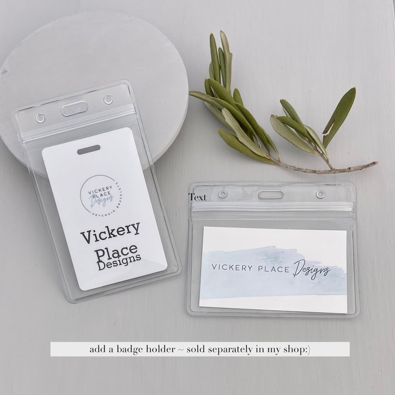 May include: Clear plastic badge holders with a white card inside. The card features the text "Vickery Place Designs" in black. A sprig of green leaves rests nearby. The text "Text" is printed on the top left of the badge holder.