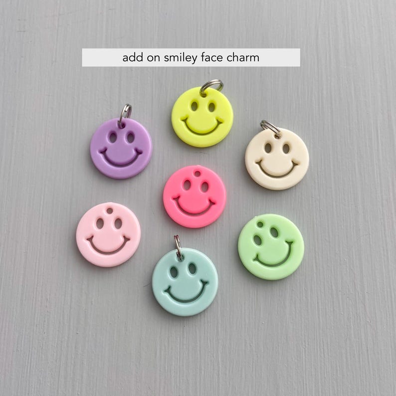 May include: A collection of colorful smiley face charms in various shades, including purple, yellow, pink, and green. Each charm has a silver-colored loop for attachment and the text "add on smiley face charm".
