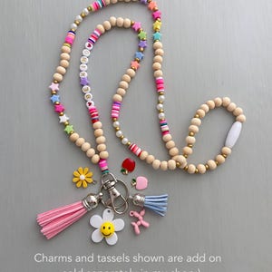 May include: A beaded lanyard with wooden beads, colorful star and letter beads, and pink and blue tassels. Additional charms include a flower, a smiling flower, an apple, and a balloon dog. The lanyard is designed to hold keys or an ID badge.