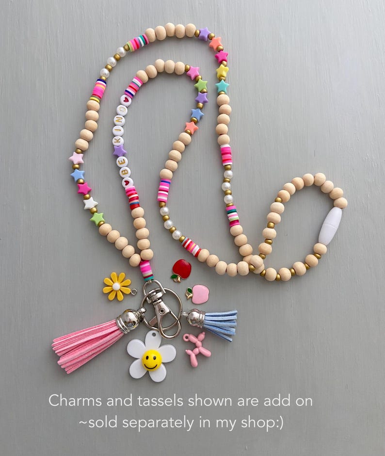 May include: A beaded lanyard with wooden beads, colorful star and letter beads, and pink and blue tassels. Additional charms include a flower, a smiling flower, an apple, and a balloon dog. The lanyard is designed to hold keys or an ID badge.