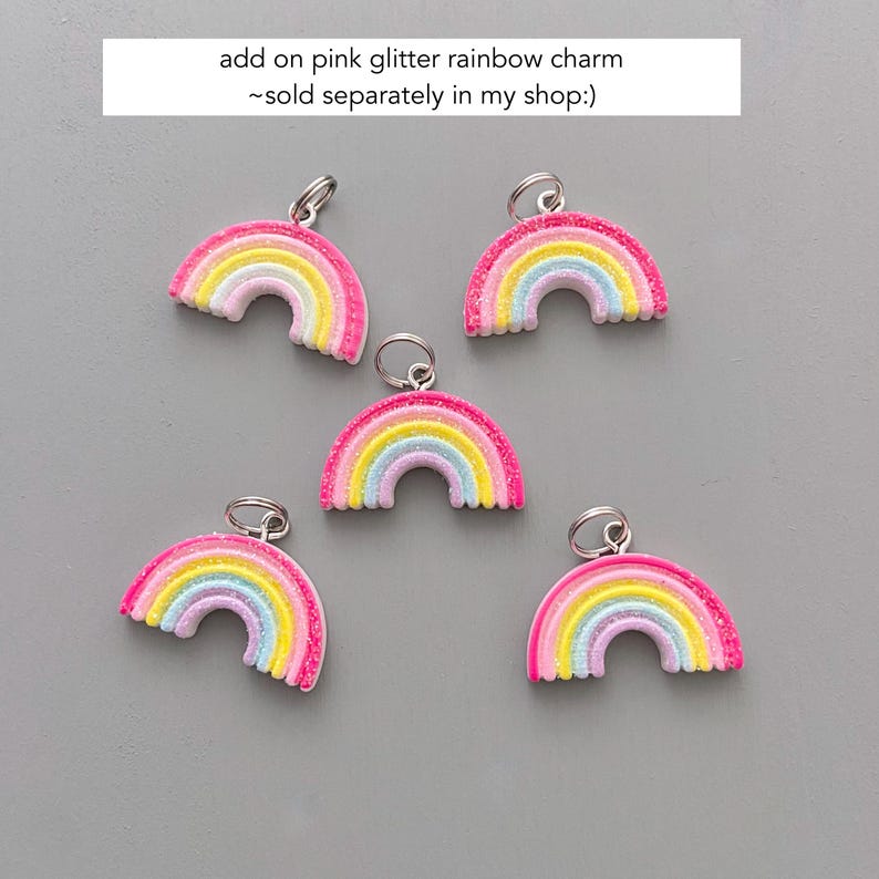 May include: Five colorful rainbow charms with pink glitter edges and silver-colored loops for attaching to jewelry or other crafts. The rainbows feature layers of yellow, pink, blue, and white. Text at the top reads "add on pink glitter rainbow charm ~ sold separately in my shop:)"