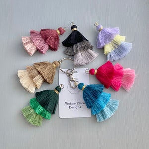 May include: A set of eight multi-colored tassel keychains with a silver key ring and a gold accent. The keychains are made of yarn and have a layered tassel design. The keychains are arranged in a circle on a light blue background. The text "Vickery Place Designs" is visible on a white card.
